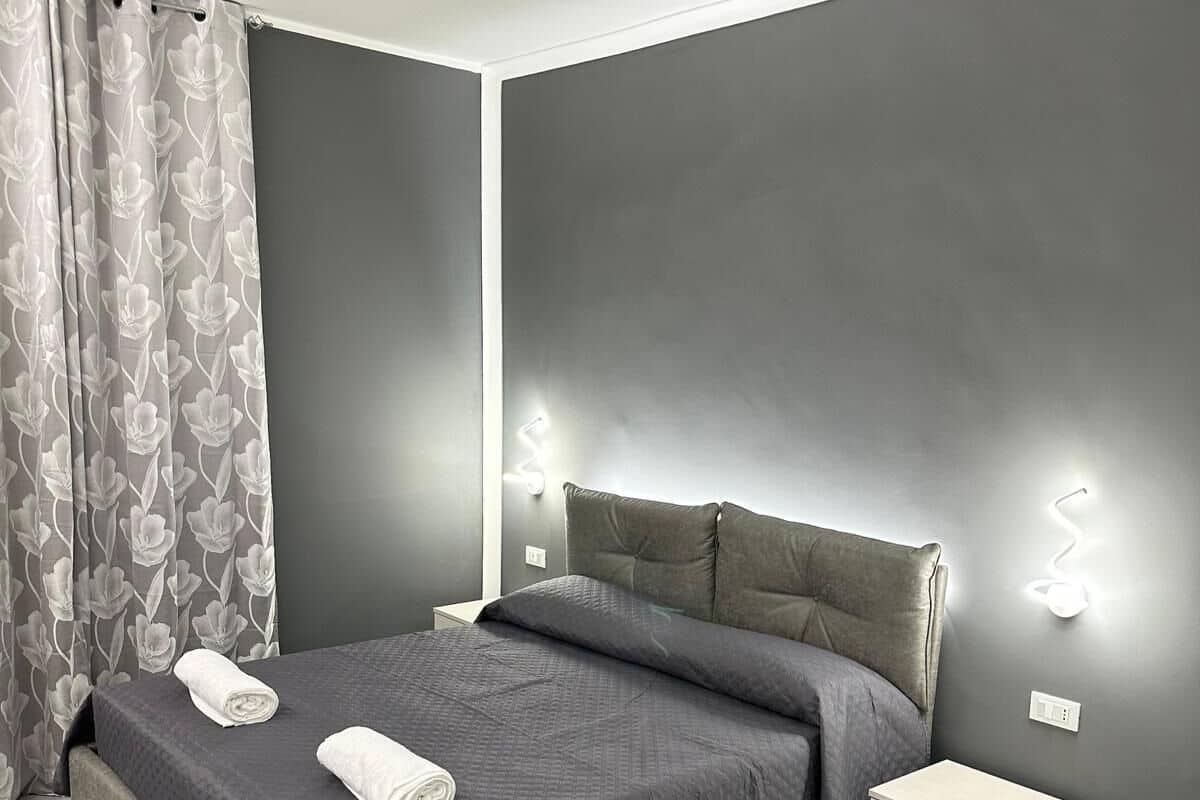 Quarto moderno com cama cinza, mesa de cabeceira de madeira e cortina cinza com padrão floral.