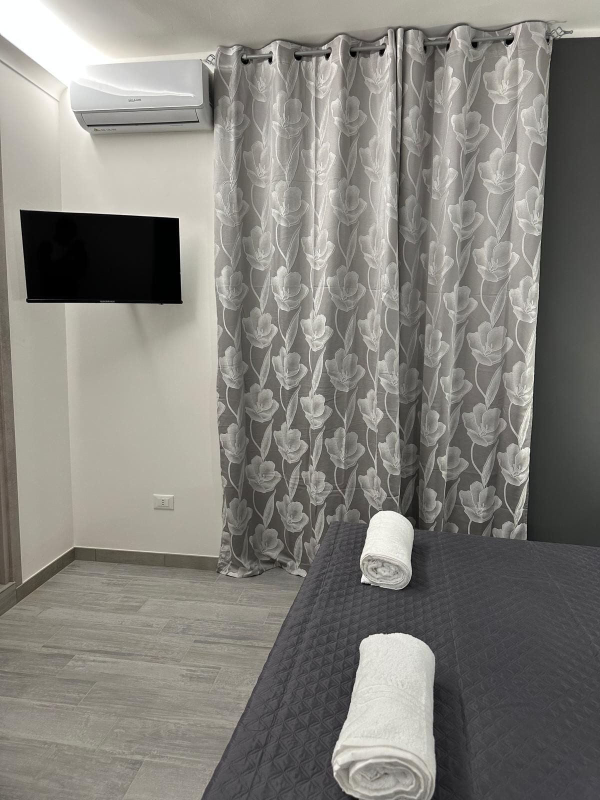 A hotel room with a bed, a TV mounted on the wall, and grey floral curtains.