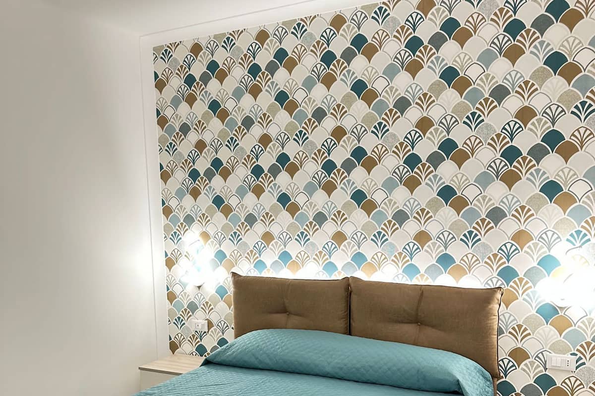 Cama bem arrumada com colcha verde-água e duas toalhas brancas, cabeceira estofada marrom e papel de parede estampado ao fundo.