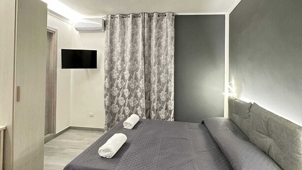 Een moderne slaapkamer met een groot bed, grijs beddengoed, een flatscreen-tv aan de muur en een grijs gordijn met een bloemenpatroon.
