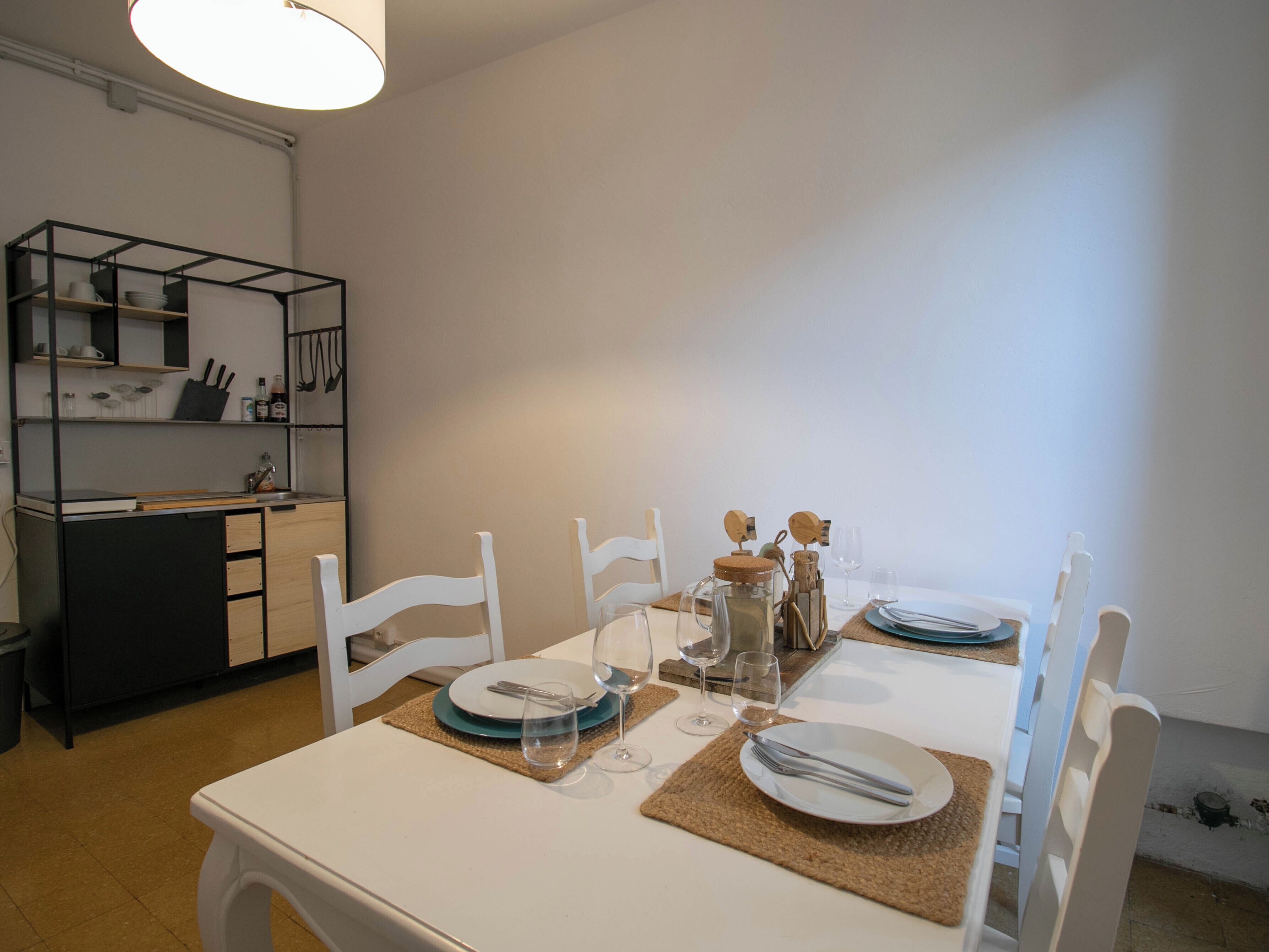 Un coin repas avec une table blanche dressée pour quatre personnes, entourée de chaises blanches. On aperçoit, à gauche, un espace cuisine avec un comptoir noir et des étagères.