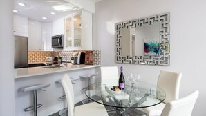 Een moderne keuken met witte kasten, een ingebouwde oven en een koffieapparaat. Er is een glazen eettafel met witte stoelen en een decoratieve spiegel erboven.