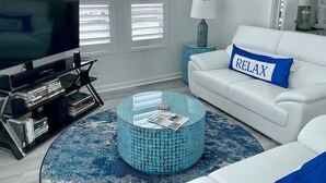 A modern living room with a TV, a white sofa with a 'RELAX' pillow, a blue patterned coffee table, and a bookshelf.