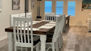 A dining room with a wooden table and white chairs, large windows with blinds, and artwork on the walls.
