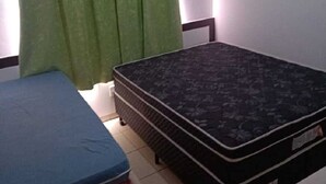 Ein Zimmer mit einem Bett, einer Matratze, einem kleinen Tisch und einem Fenster mit grünen Vorhängen.