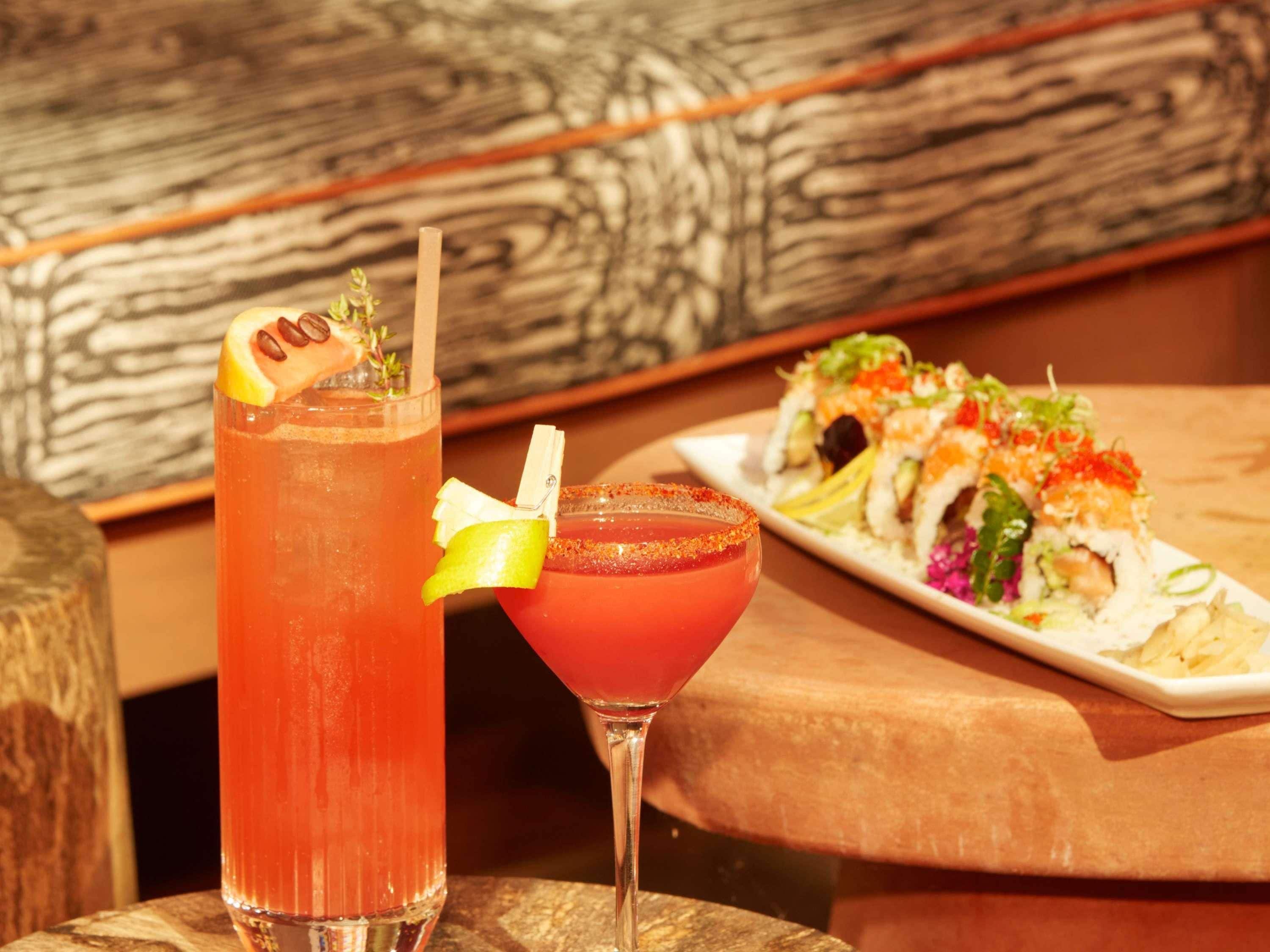 Two cocktails on a wooden table with a sushi dish in the background.