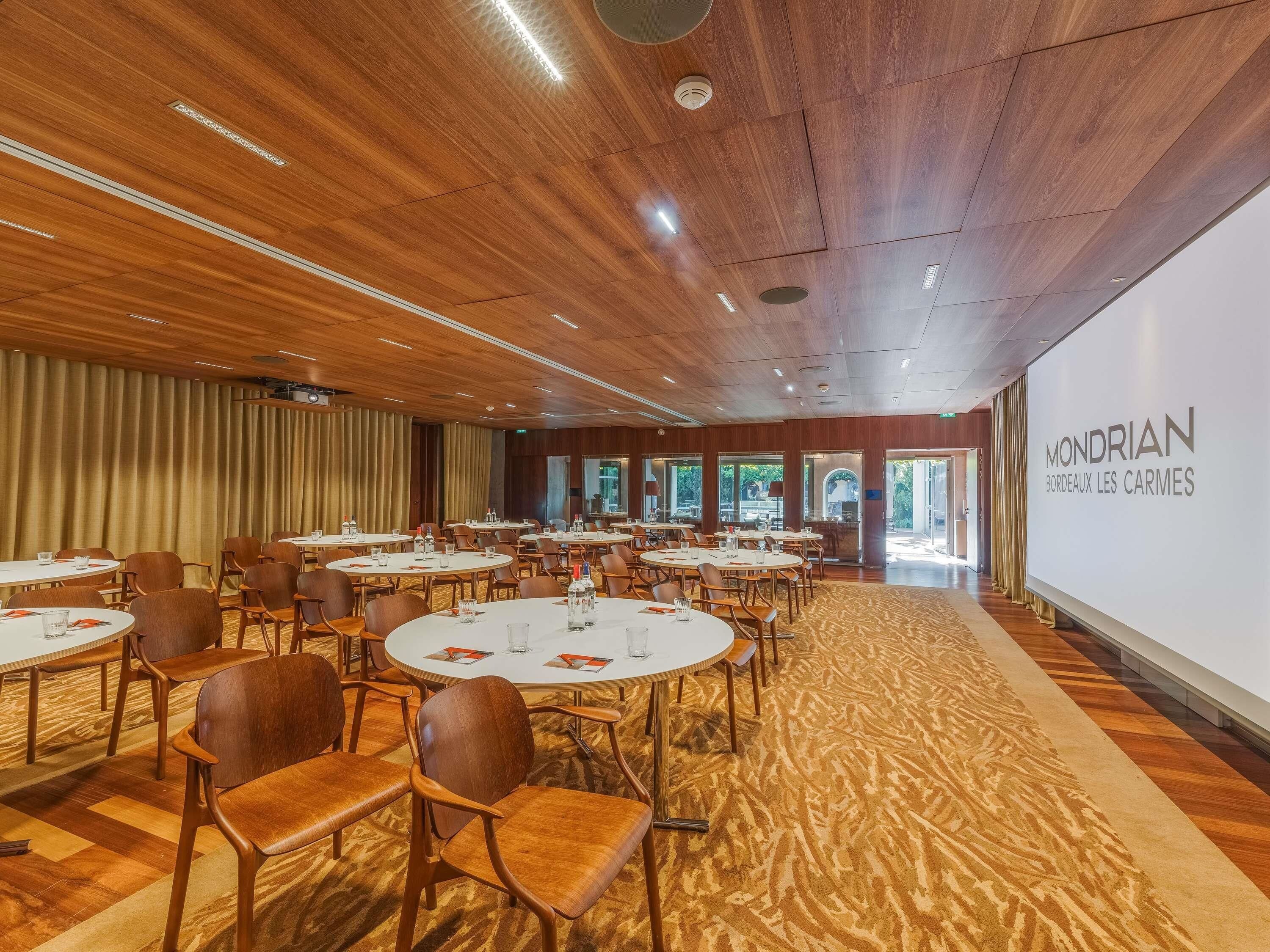 A conference room with round tables and chairs, a wooden ceiling, and a large screen displaying 'MONDRIAN BORDOEX LES CARMES'.