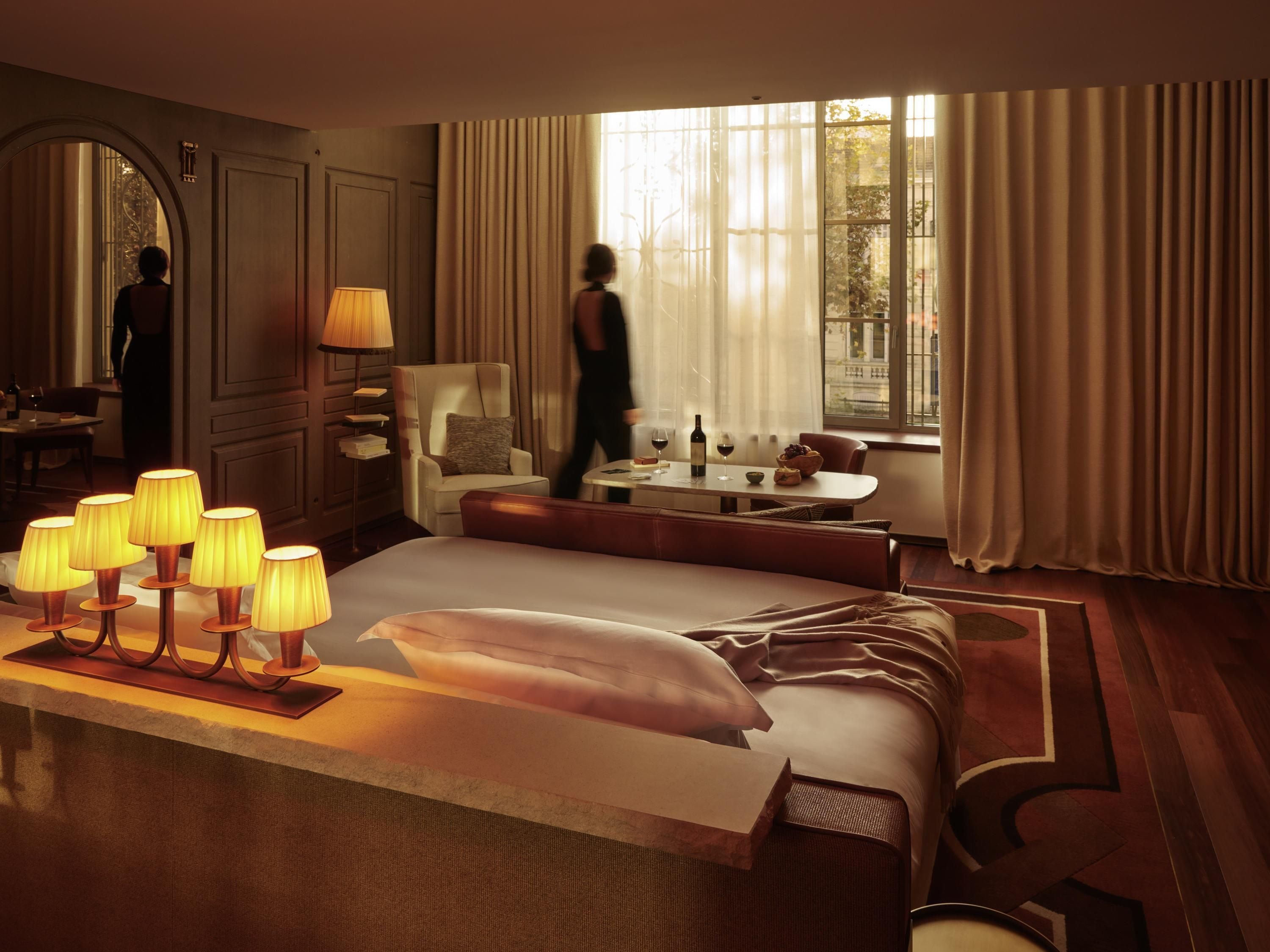 Une chambre d’hôtel dotée d’un grand lit, d’un coin salon avec une table et des chaises, d’une fenêtre avec des rideaux et d’un lampadaire muni de plusieurs lampes.