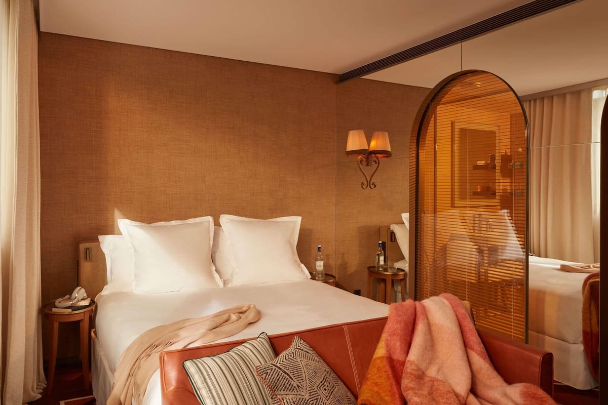 A hotel room with a large bed, a bedside table with a lamp, a mirror, and a view of another room.