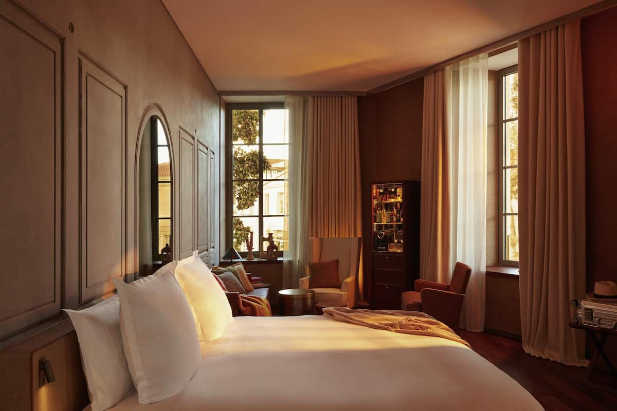 A hotel room with a large bed, a seating area with chairs, a small table, and a cabinet with bottles.