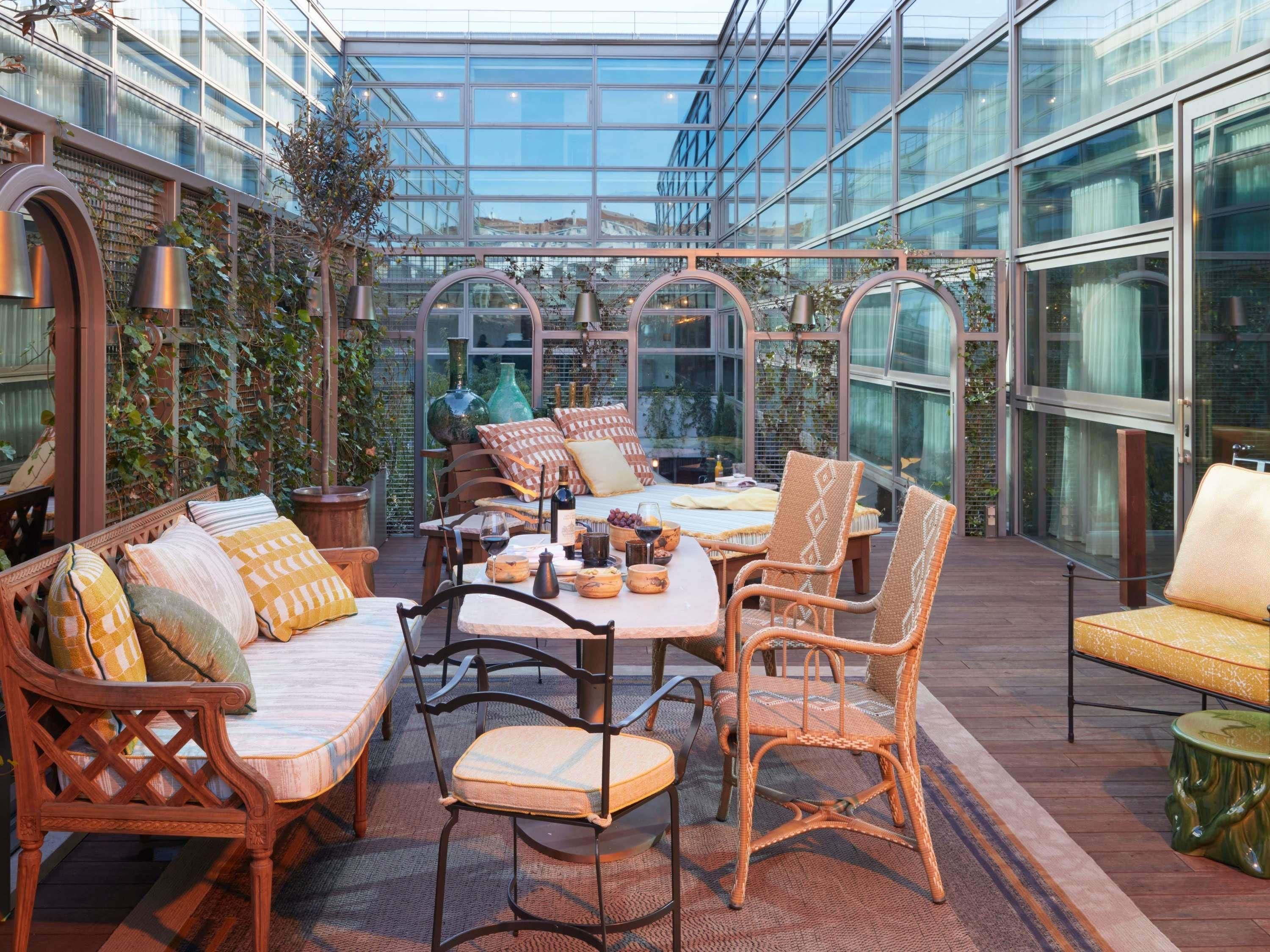 Un porche con paredes de vidrio, muebles de mimbre, una mesa preparada con comida y bebidas, y vista al paisaje urbano.