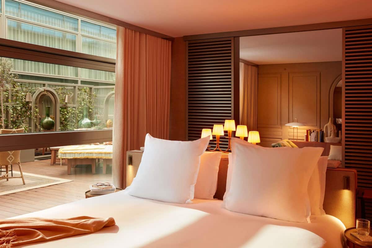 A hotel room with a large bed, bedside lamps, a view of greenery outside, and a dining area with a table and chairs.