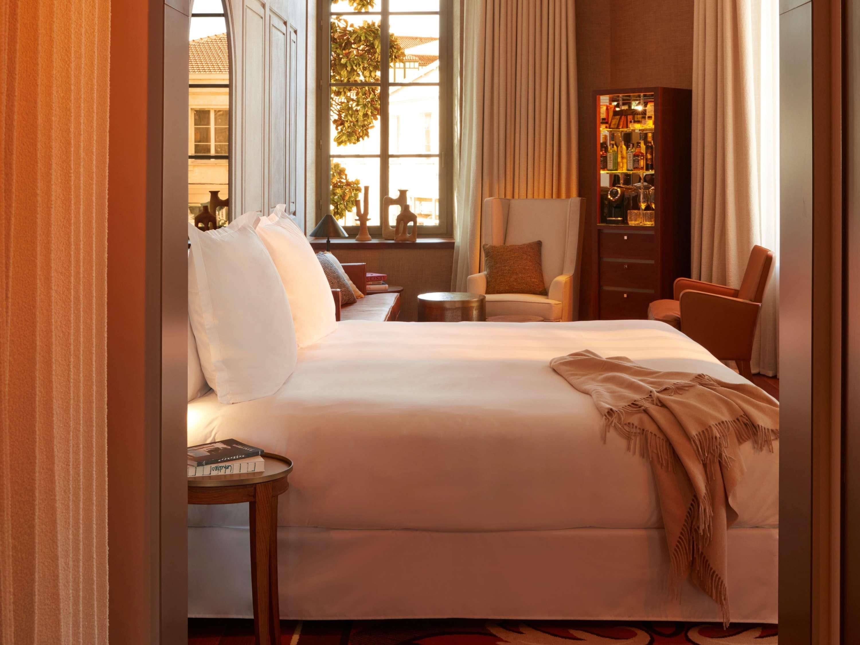 Habitación de hotel con una cama grande, un área de estar con una silla y una mesita, una estantería y una ventana con cortinas.