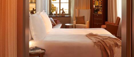 Habitación de hotel con una cama grande, un área de estar con una silla y una mesita, una estantería y una ventana con cortinas.