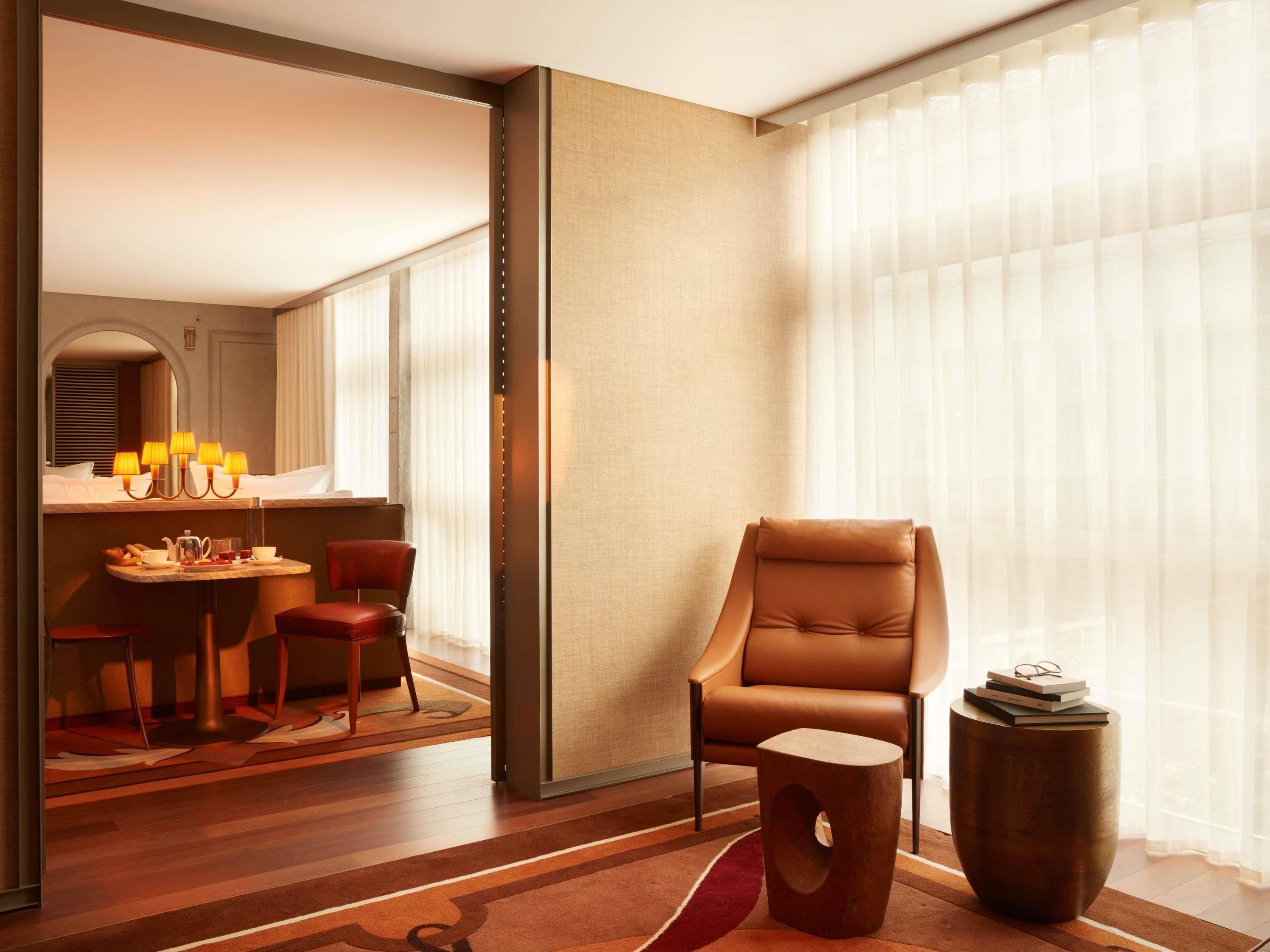 A modern hotel room with a dining area, a leather armchair, a round side table, and a large window with sheer curtains.