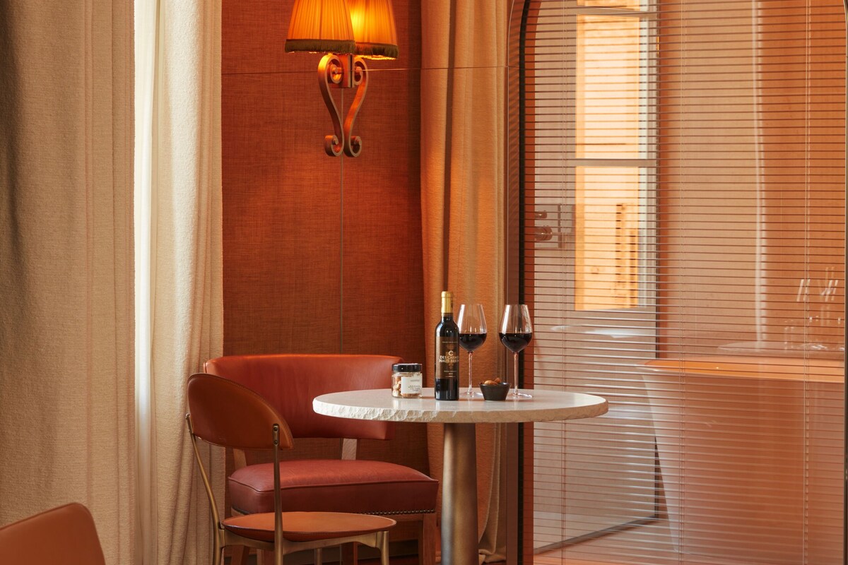 Habitación de hotel con una mesa redonda, dos sillas, una lámpara y vistas a trav és de un gran ventanal con persianas.