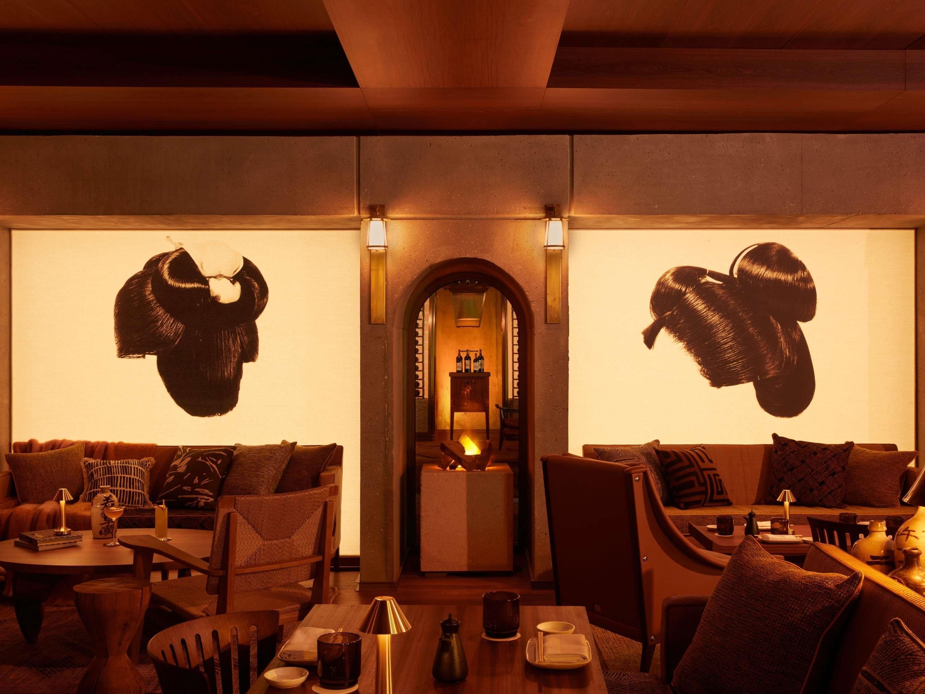 A cozy restaurant interior with two sofas, a table, and chairs. There are wall-mounted lamps and a small bar area visible through an archway.