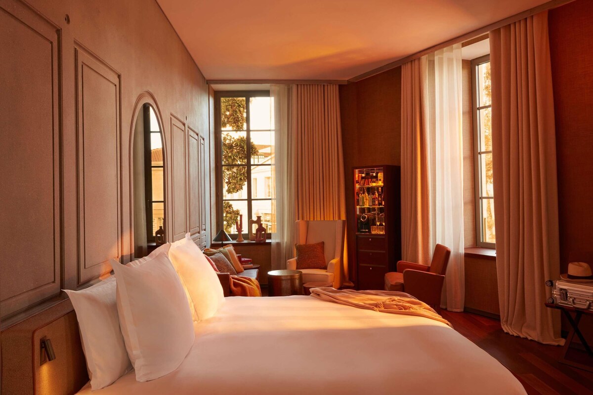 Una habitación de hotel con una cama grande, zona de estar con sillas, una mesita y un mini bar.