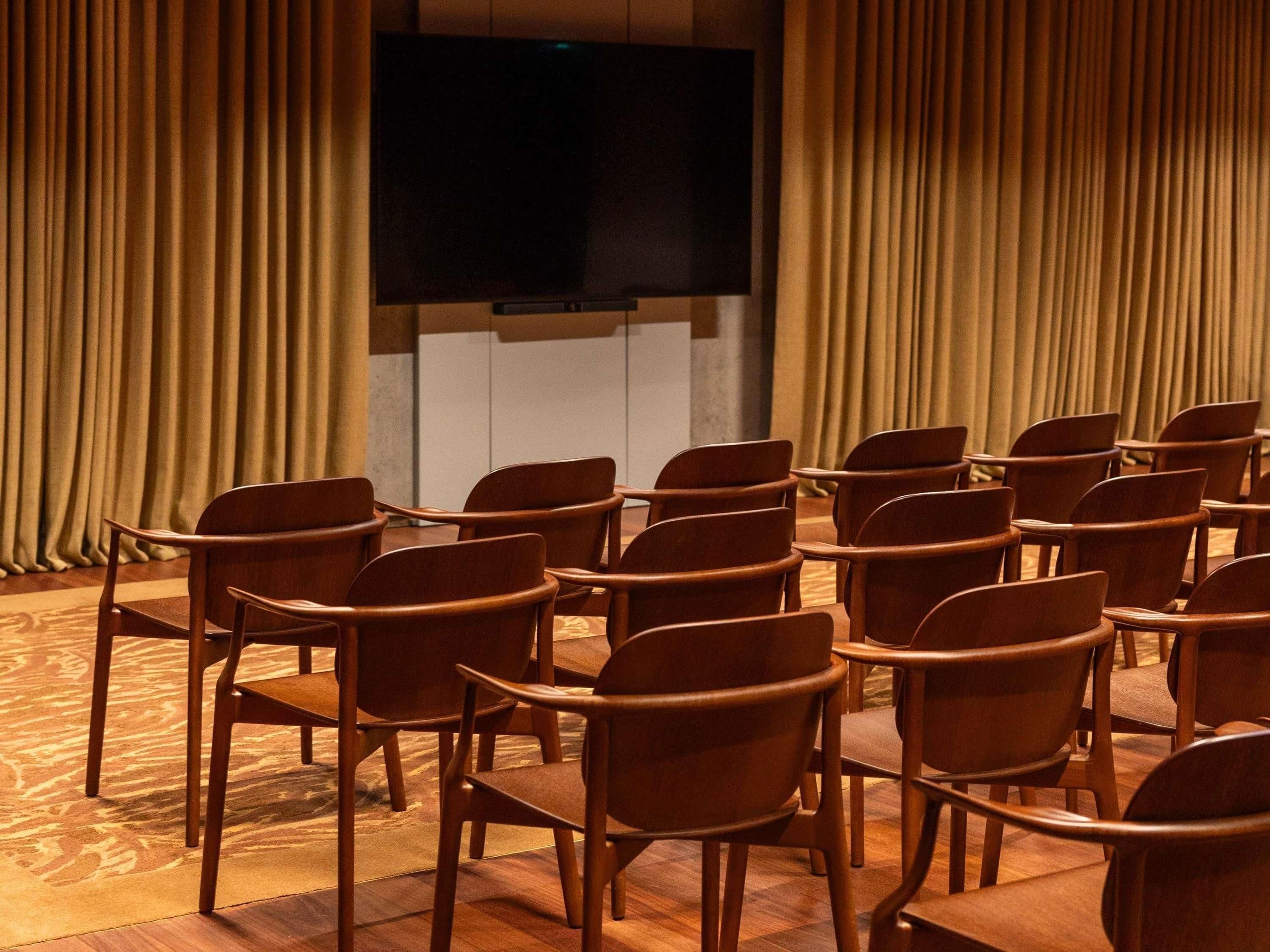 Una sala de conferències amb files de cadires orientades cap a una pantalla gran, cortines als laterals i una catifes amb disseny.