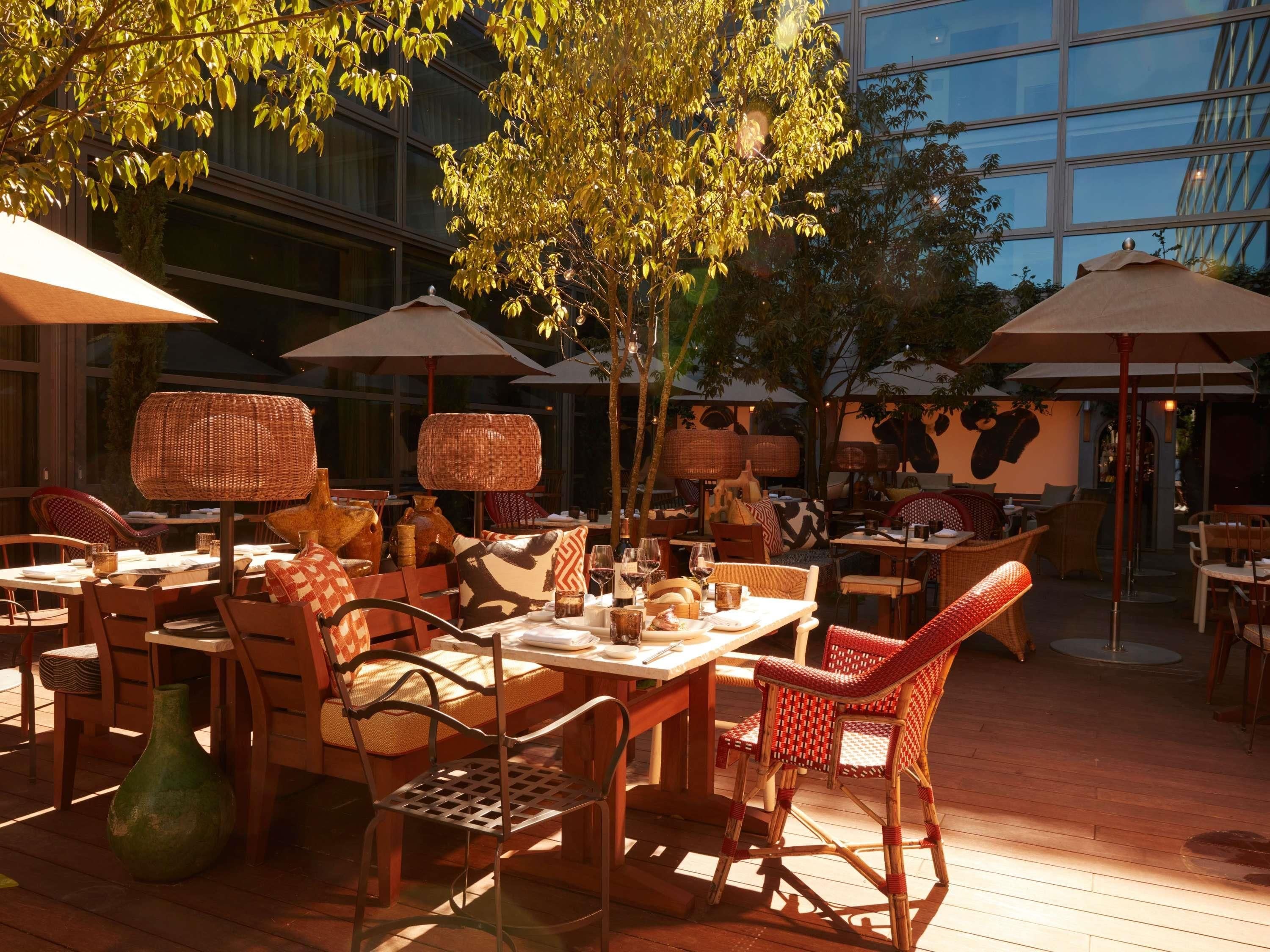 Un área de comedor al aire libre con mobiliario de mimbre, sombrillas y vistas a un edificio moderno.