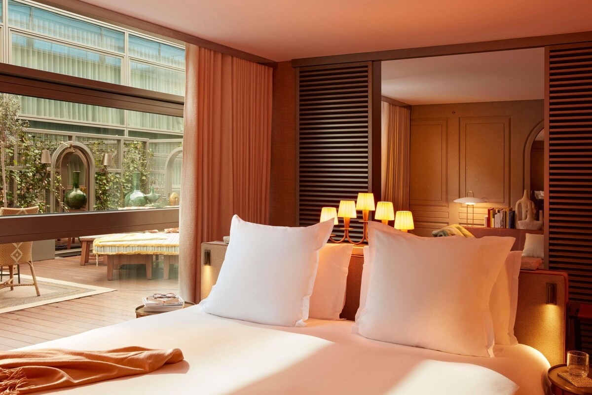 A hotel room with a large bed, bedside lamps, a view of greenery outside, and a dining area with a table and chairs.