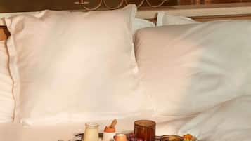 A bed with white linens, a bedside table with a glass of milk, a bottle of lotion, a jar of jam, and a plate with pastries.