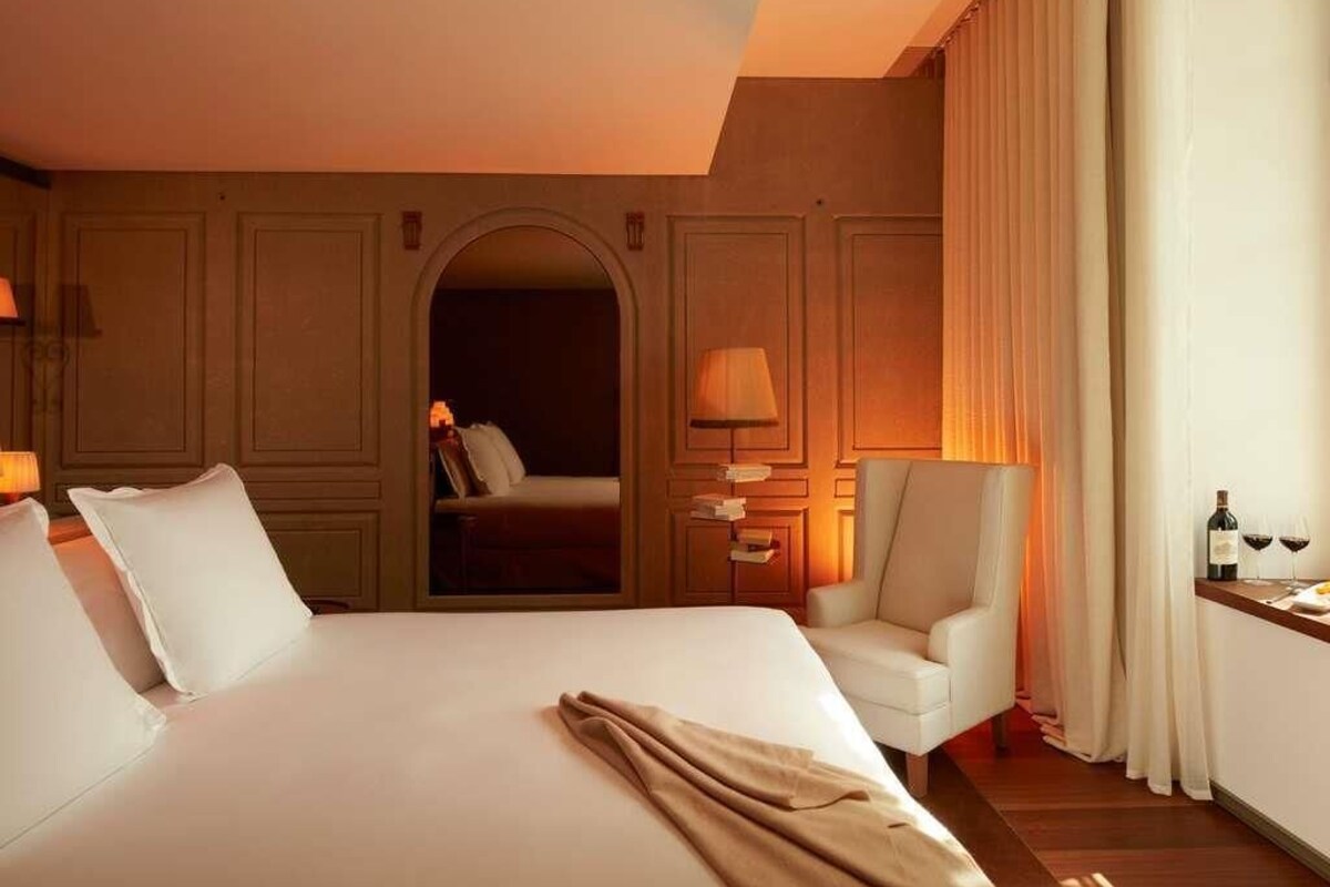 Habitación de hotel con una cama grande, zona de estar con una silla, escritorio con lámpara y vistas a través de un arco.