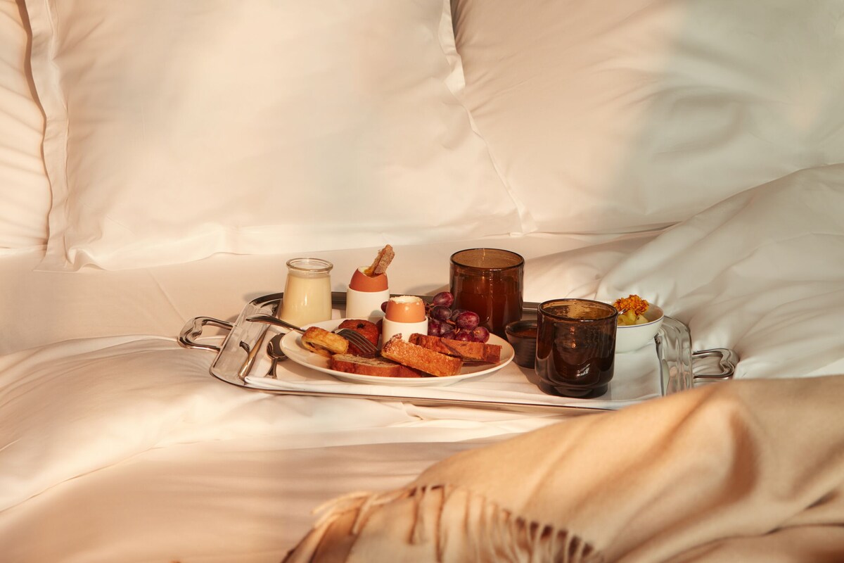 Una habitación de hotel con una cama bien hecha, una bandeja con alimentos para el desayuno y varias lámparas de mesa.