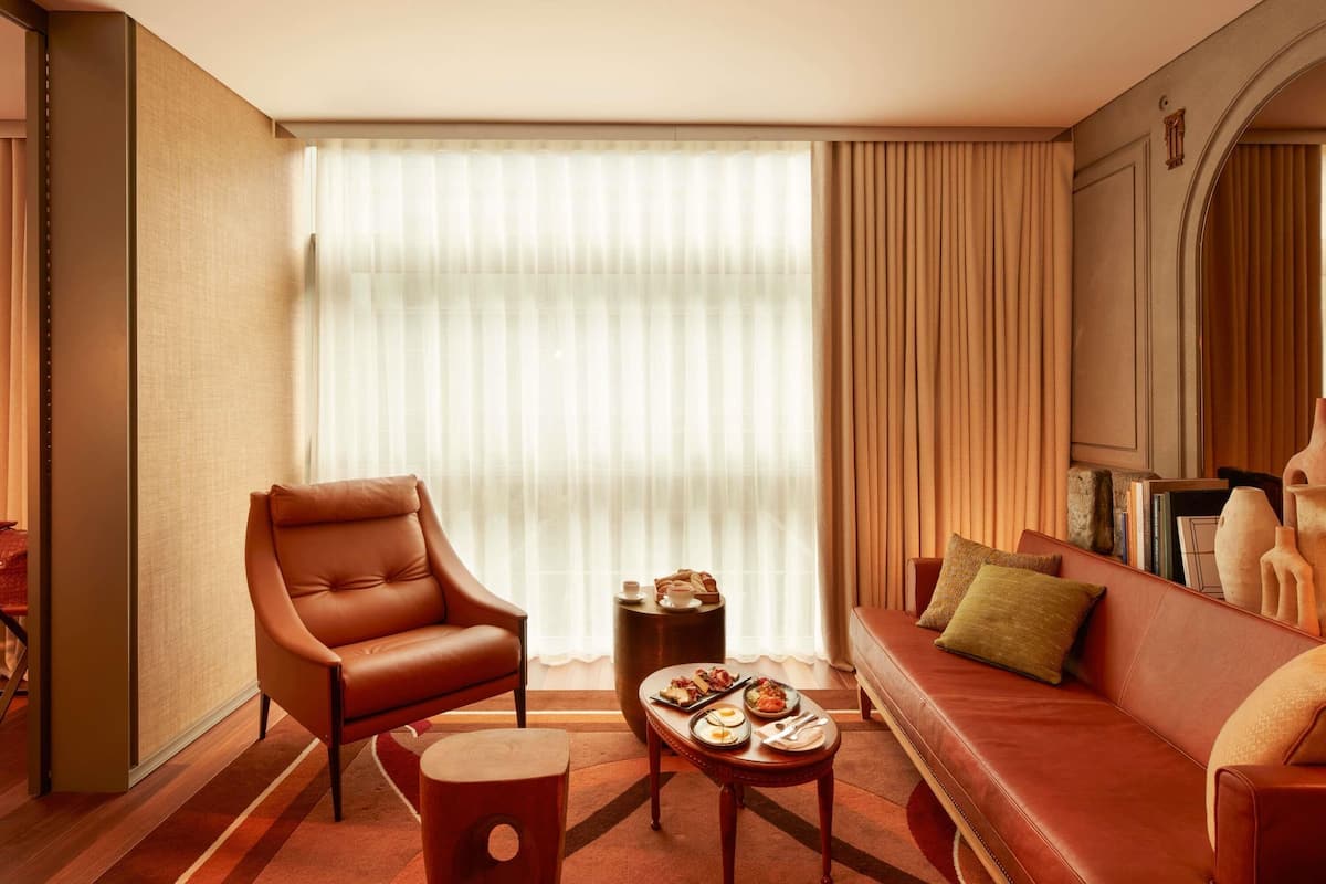 A modern living room with a leather sofa, a leather armchair, a small round table with a tray of food, and a large window with sheer curtains.