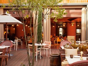 An outdoor dining area with wooden tables and chairs, surrounded by trees and columns. - Mondrian Bordeaux Les Carmes (Bordeaux)