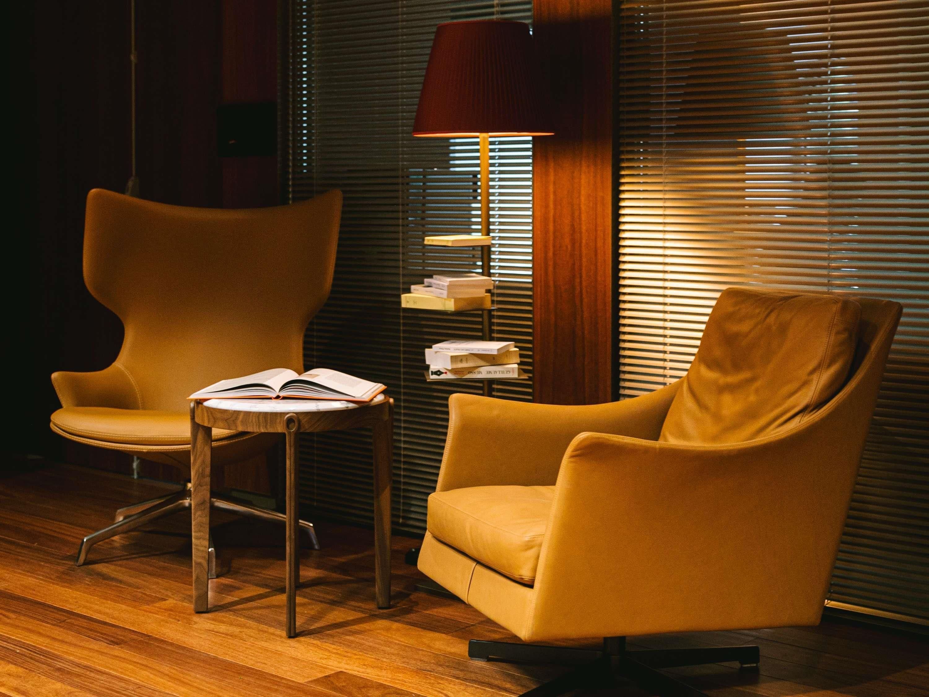 Een kamer met twee fauteuils, een houten bijzettafeltje met een open boek, een lamp en een boekenkast tegen een wand met jaloezieën.