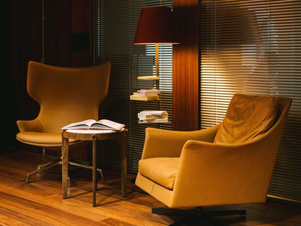 Una habitación con dos sillones, una mesita de madera con un libro abierto, una lámpara y una estantería contra una pared con persianas.