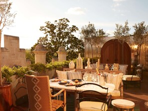 Un área de comedor al aire libre con una mesa dispuesta para dos personas, rodeada de sillas de mimbre y un banco acolchado.