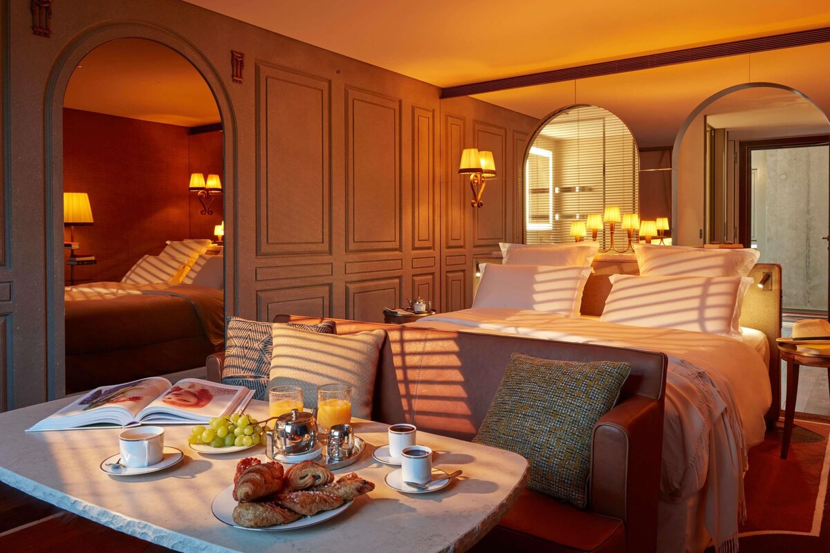 A hotel room with a large bed, a sitting area with a table set for breakfast, and a view through the window.