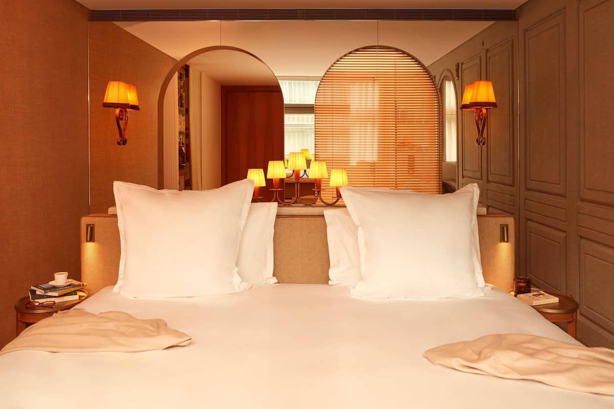 A hotel room with a large bed, two bedside lamps, a nightstand with a book and glasses, and a view of the hallway.