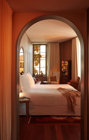 A hotel room with a large bed, a sitting area with a chair and a small table, a bookshelf, and a window with curtains. - Mondrian Bordeaux Les Carmes (Bordeaux)