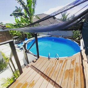 Une piscine extérieure dotée d’une terrasse en bois, entourée de plantes tropicales et agrémentée d’un espace ombragé.