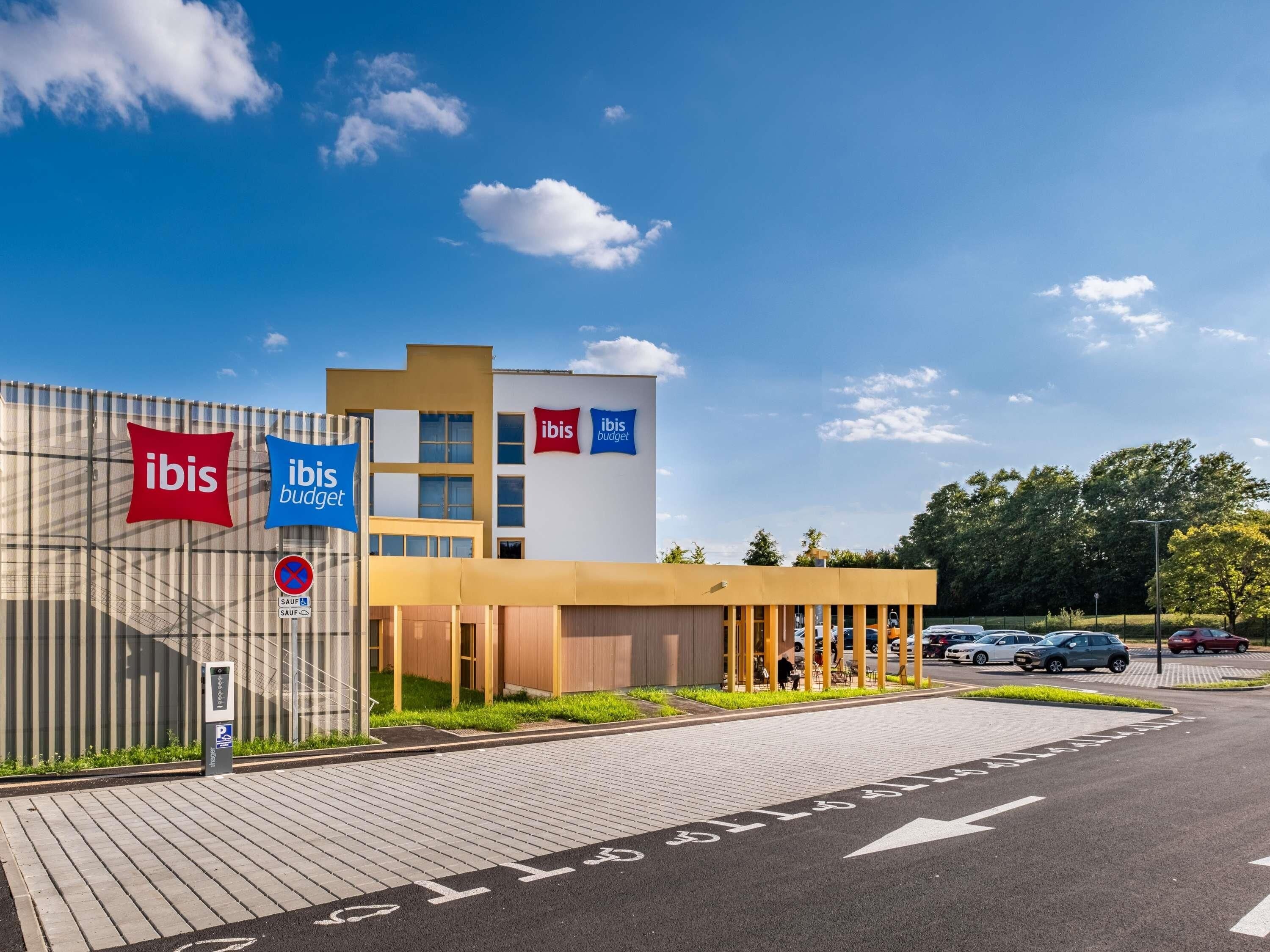 A modern hotel with 'ibis' and 'ibis budget' signage, a parking area, and a clear blue sky.
