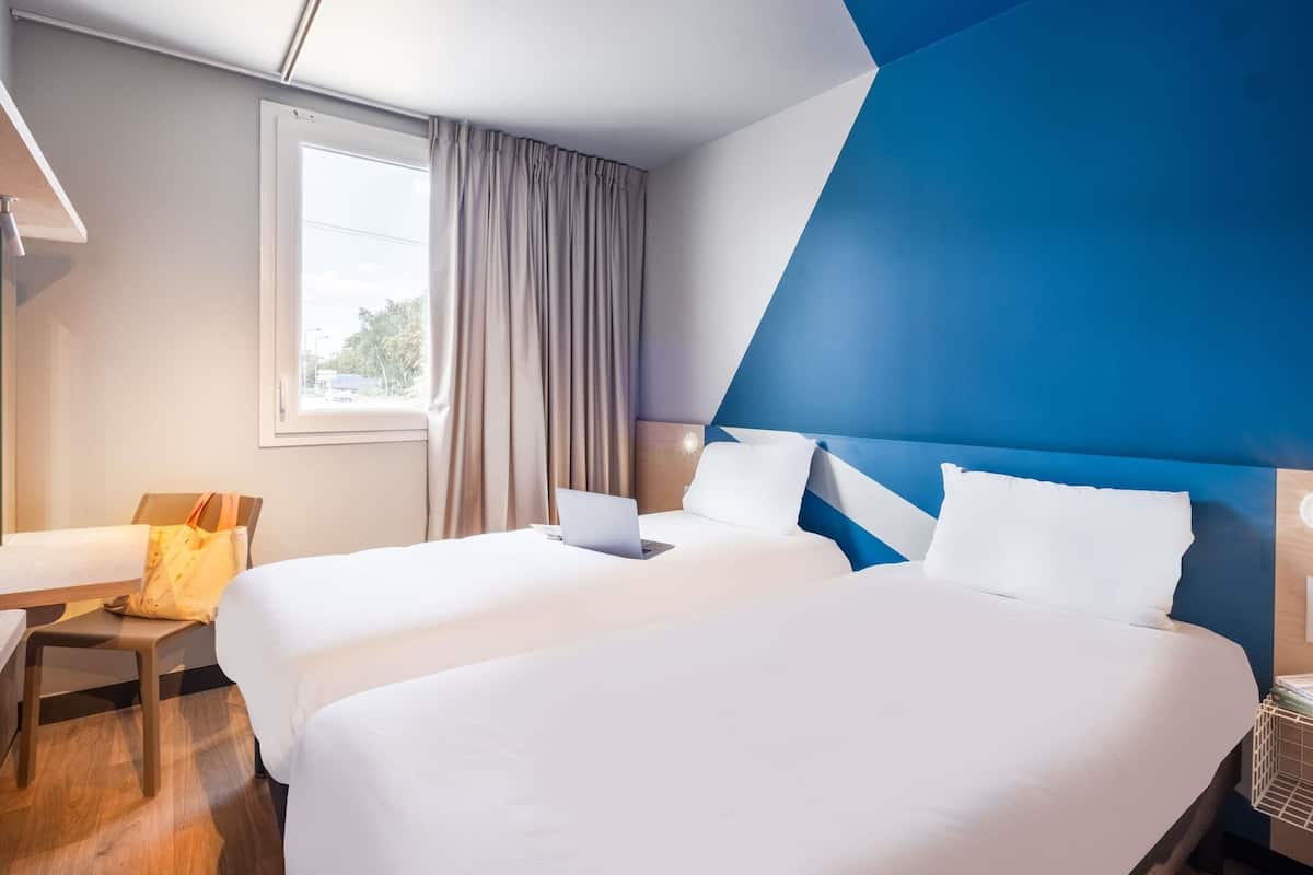 Une chambre d’hôtel avec deux lits, un bureau, une chaise, une fenêtre et un mur d’accent bleu.