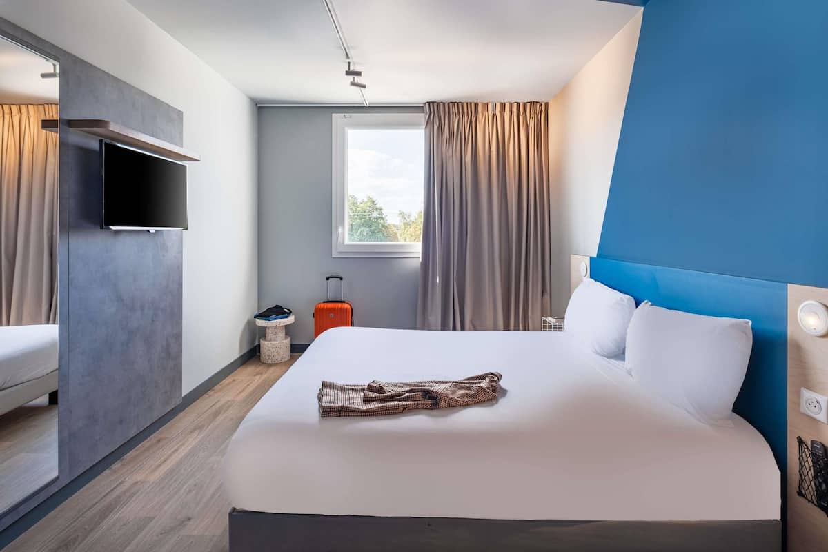 Une chambre d’hôtel moderne équipée d’un lit, d’une télévision, d ’une fenêtre avec des rideaux et d’une valise.