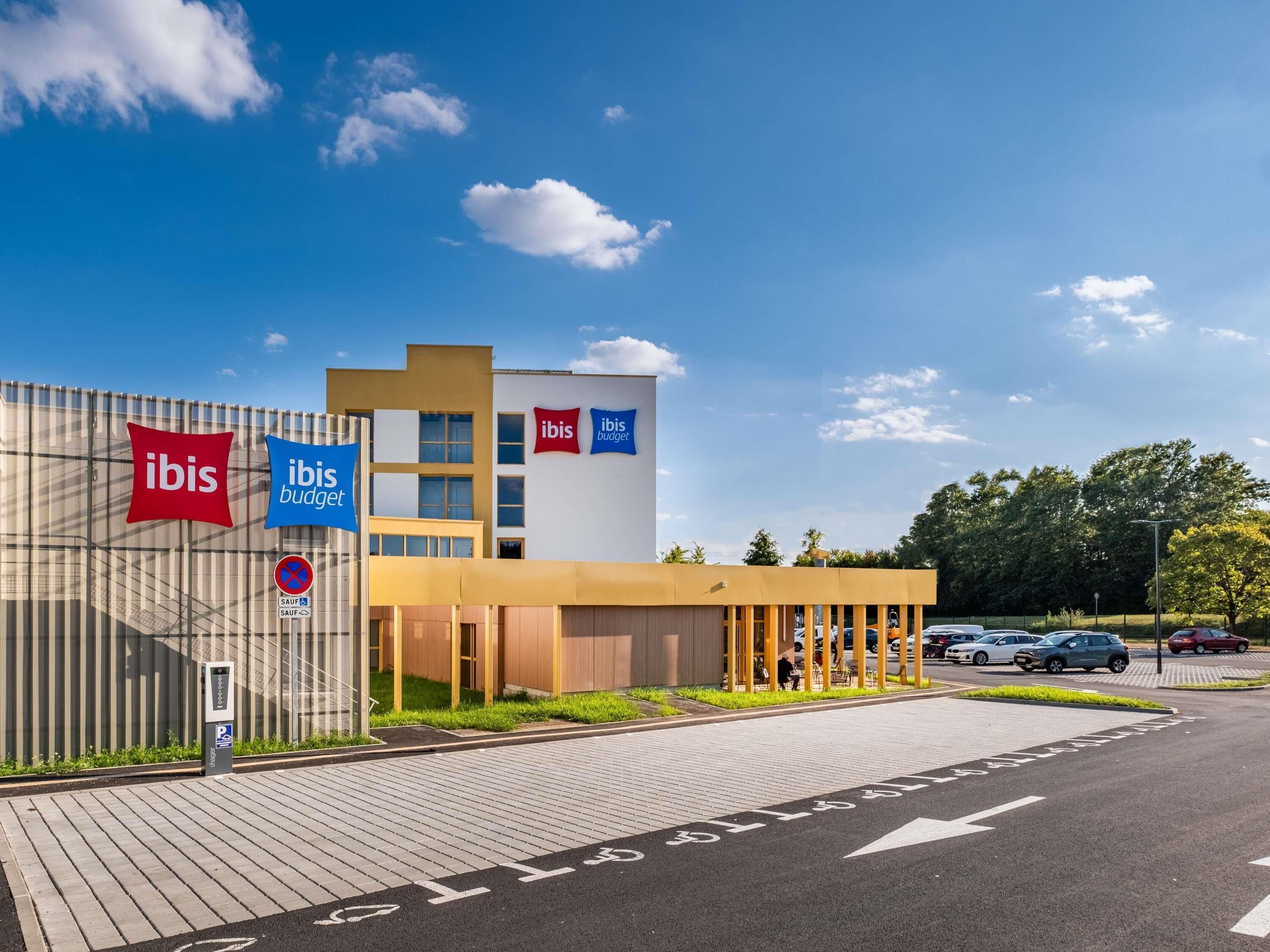 Moderni hotelli, jossa on näkyvillä »ibis»- ja »ibis budget» -kyltit, pysäköintialue ja kirkas, sininen taivas.