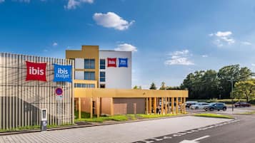 Moderni hotelli, jossa on näkyvillä »ibis»- ja »ibis budget» -kyltit, pysäköintialue ja kirkas, sininen taivas.