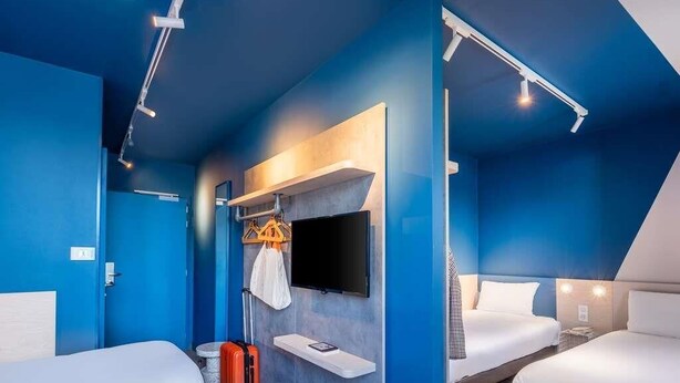 A hotel room with two beds, a TV mounted on the wall, and a suitcase on the floor. - Ibis Budget Nantes Nord Treillières (Treillieres)