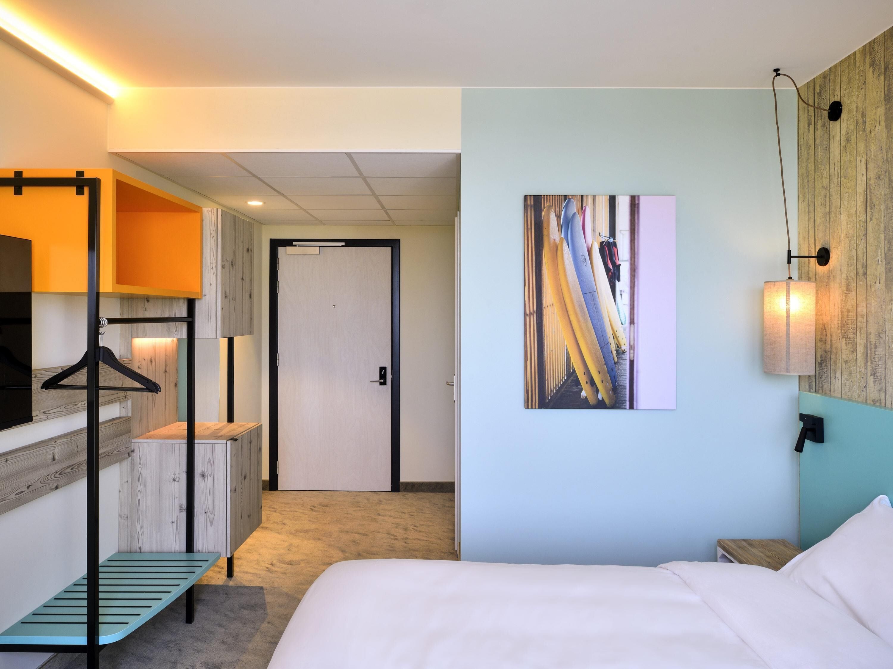 Hotelkamer met een bed, een bankje, een kapstok, een deur en een wandlamp.