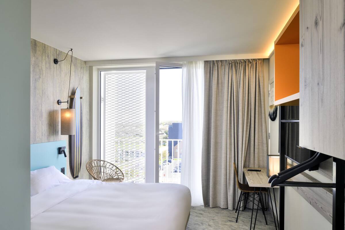 Une chambre d’hôtel avec un lit, un bureau et une chaise. Il y a une fenêtre avec des rideaux et une vue sur les bâtiments environnants.