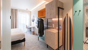 A hotel room with a bed, a desk with a chair, a small table, a bench, a surfboard, and a wardrobe. - Ibis Styles Bredene  (Bredene)