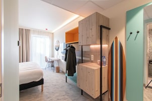 Habitación de hotel con una cama, un escritorio con una silla, una mesita, una banca, una tabla de surf y un armario.