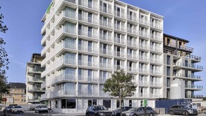 A multi-story hotel with balconies, a green logo, and a parking area with cars. - Ibis Styles Bredene  (Bredene)