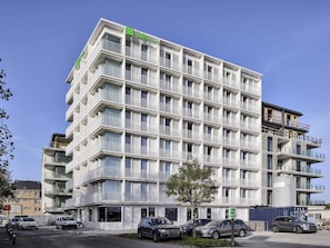 A multi-story hotel with balconies, a green logo, and a parking area with cars. - Ibis Styles Bredene  (Bredene)