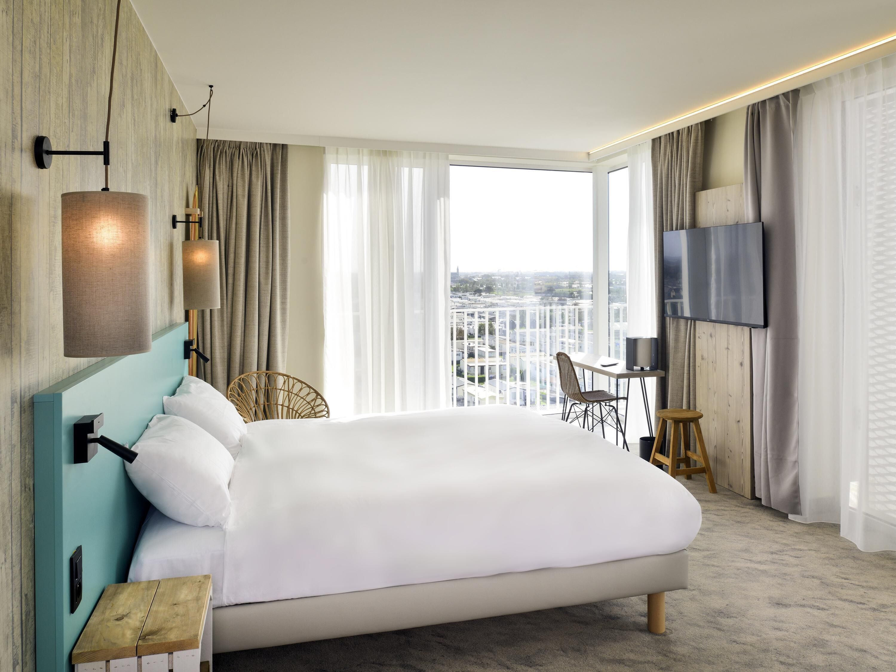 A modern hotel room with a large bed, a desk with a chair, a TV, and a view of the city.